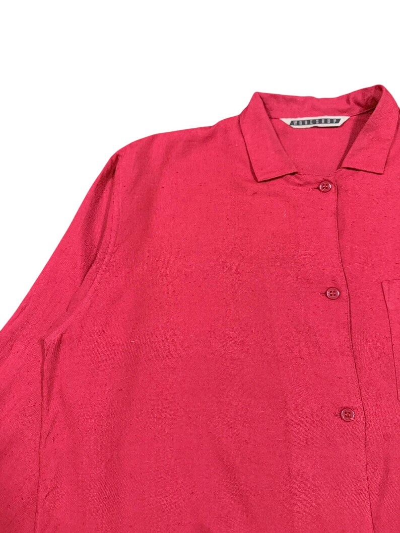 Puede incluir: Primer plano de una camisa roja con cuello, cierre de botones y bolsillo. La camisa tiene mangas largas y una superficie texturizada. El nombre de la marca es visible en el cuello.
