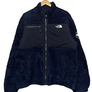 May include: A dark navy blue fleece jacket with a high collar and a full-length front zipper. The garment features a smooth black chest panel with a white The North Face logo, a zippered chest pocket, two lower pockets, and a circular patch on the sleeve.