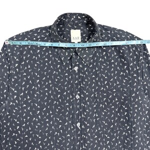 May include: A dark gray button-down shirt with a white repeating floral pattern and vertical textured stripes. A measuring tape across the shoulders shows a width of approximately 19 inches. The shirt features a collar, a front pocket, and white buttons.
