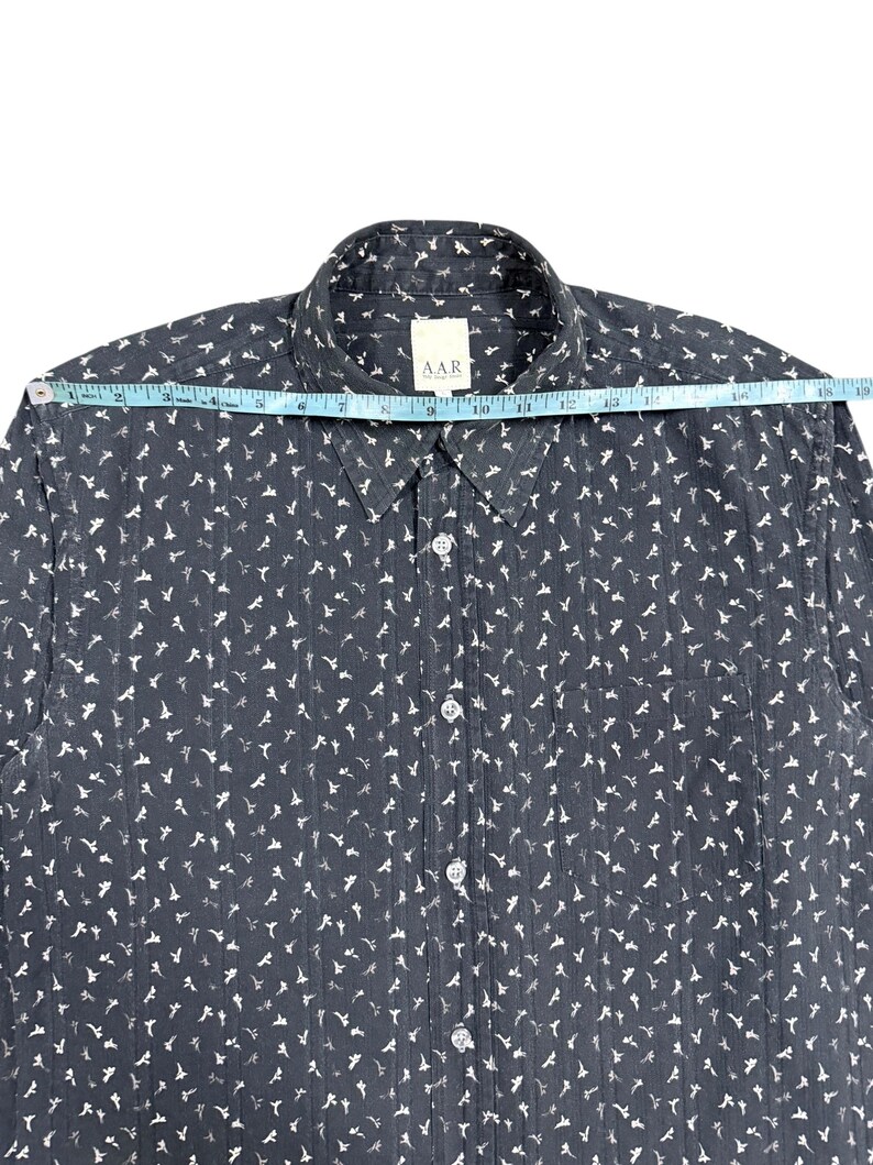 May include: A dark gray button-down shirt with a white repeating floral pattern and vertical textured stripes. A measuring tape across the shoulders shows a width of approximately 19 inches. The shirt features a collar, a front pocket, and white buttons.