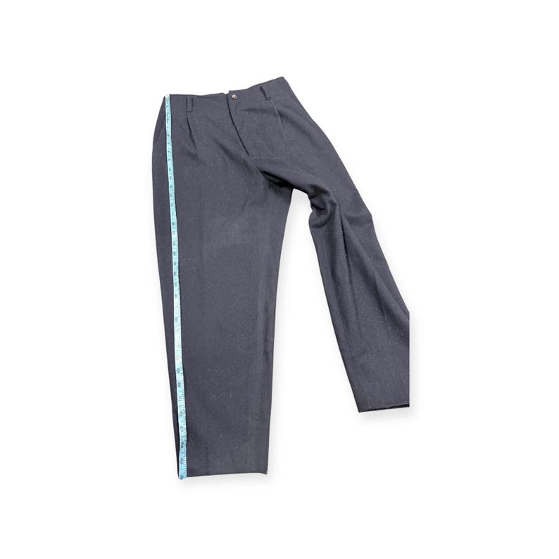 Puede incluir: Pantalones de color gris carb&oacute;n oscuro con frente plisado, cierre de bot&oacute;n y trabillas para cintur&oacute;n. Una cinta m&eacute;trica azul claro est&aacute; colocada a lo largo de la costura lateral, indicando una longitud de unos 107 cm desde la cintura al dobladillo.