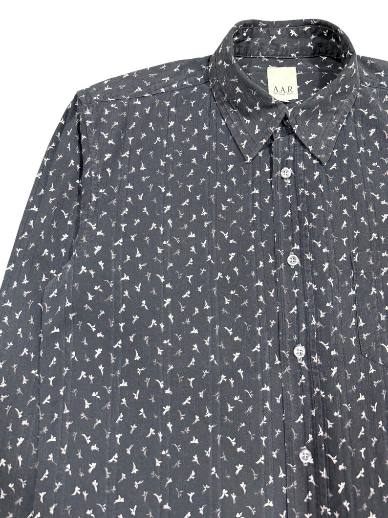 May include: A dark gray long-sleeve button-down shirt featuring a repeating white floral pattern and vertical textured stripes. The shirt has a standard collar, a front button placket with clear buttons, and a chest pocket. A label reads A.A.R.