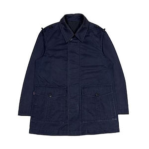 May include: A dark navy blue utility jacket featuring a classic collar, a concealed front button placket, and two large rectangular patch pockets on the lower front with buttoned flaps. The jacket includes shoulder epaulets with small buttons.