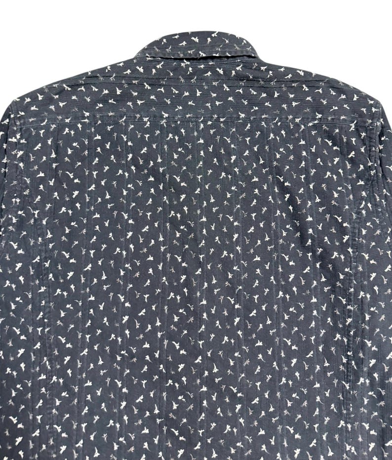 May include: A dark charcoal gray long-sleeve shirt featuring a repeating white floral pattern and vertical textured stripes. The garment includes a standard collar, a back yoke, and a button-down construction with a consistent all-over print.