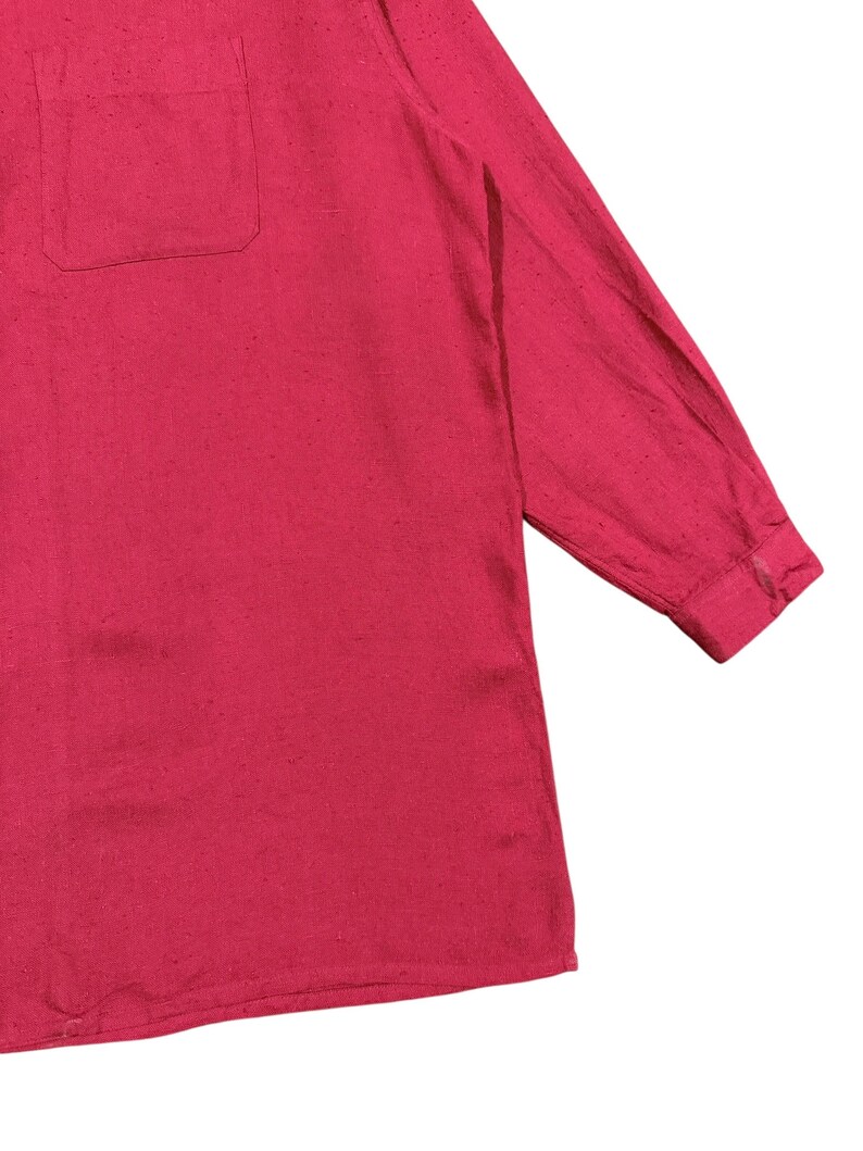 Puede incluir: Una camisa roja de manga larga con un bolsillo en el pecho. La camisa tiene un pu&ntilde;o abotonado y una tela ligeramente texturizada. La prenda es de color liso y parece estar hecha de un material ligero.