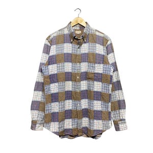 Op de afbeelding: Een overhemd met lange mouwen en knoopsluiting, voorzien van een patchwork-ruitpatroon in bruin-, wit- en blauwtinten. Het overhemd heeft een puntige kraag, een enkele borstzak en een volledige knoopsluiting aan de voorzijde, getoond op een kledinghanger.