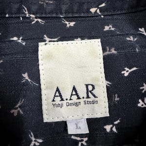 May include: A dark navy blue shirt featuring a small white abstract floral pattern. A rectangular white fabric label is stitched to the inner collar, displaying the text A.A.R Yohji Design Studio in black, with a smaller white tag below marked L.