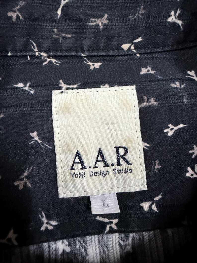 May include: A dark navy blue shirt featuring a small white abstract floral pattern. A rectangular white fabric label is stitched to the inner collar, displaying the text A.A.R Yohji Design Studio in black, with a smaller white tag below marked L.