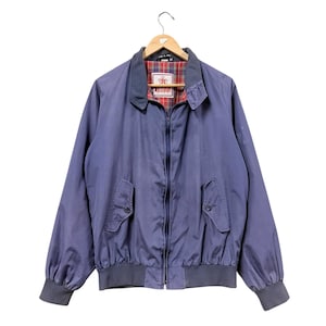 Könnte beinhalten: Eine blaue Jacke mit Reißverschluss, klassischem Kragen, langen Ärmeln mit gerippten Bündchen und einem gerippten Saum. Die Jacke verfügt über zwei Vordertaschen mit geknöpften Klappen und ein rot-grün-blau kariertes Innenfutter. Ein weißes Etikett ist sichtbar.