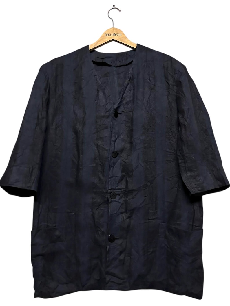 May include: A dark navy blue button-down shirt with a textured, crinkled fabric finish. The shirt features a V-neckline, elbow-length sleeves, and two lower front pockets. It is displayed on a wooden hanger labeled French Connection.