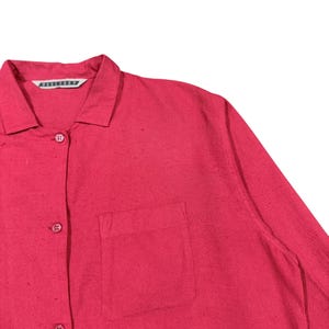 Puede incluir: Primer plano de una camisa roja brillante con botones y bolsillo en el pecho. La camisa tiene cuello y botones en la parte delantera. La tela tiene una textura sutil.