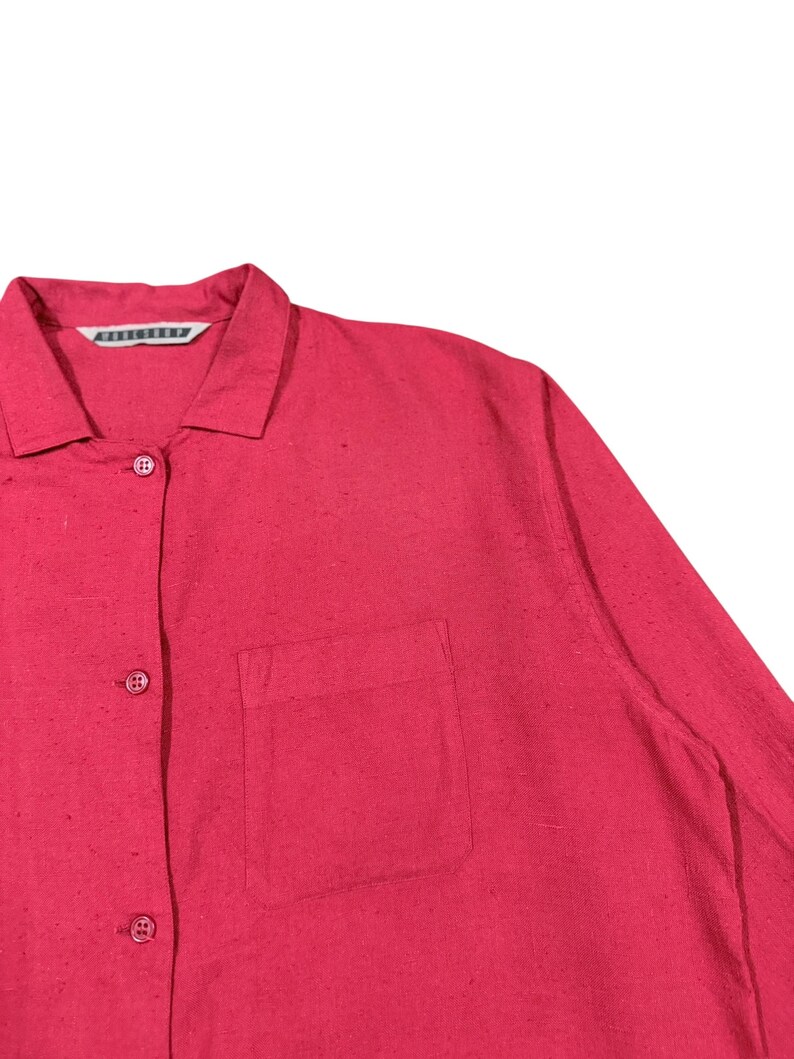Puede incluir: Primer plano de una camisa roja brillante con botones y bolsillo en el pecho. La camisa tiene cuello y botones en la parte delantera. La tela tiene una textura sutil.
