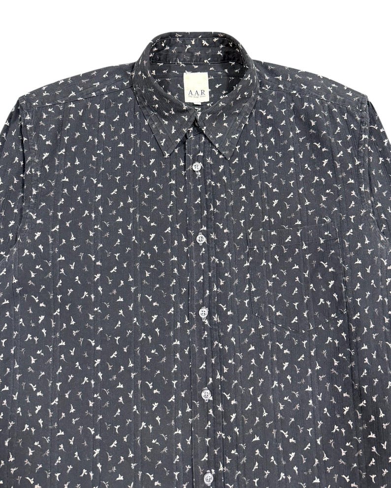 May include: A dark charcoal gray long-sleeve button-down shirt featuring a repeating white floral pattern and vertical textured stripes. The shirt includes a standard collar, a front chest pocket, and a white label with the text A.A.R.