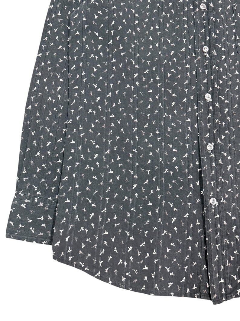 May include: A long-sleeved button-down shirt in a dark charcoal gray color featuring a repeating pattern of small white floral shapes and vertical textured stripes. The garment includes a standard collar, a front button placket, and cuffs.