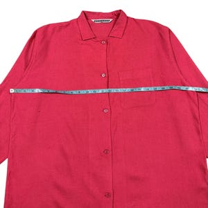 Puede incluir: Una camisa roja brillante con botones, un bolsillo en el pecho y mangas largas. La camisa se mide con una cinta m&eacute;trica azul. La camisa tiene cuello y el nombre de la marca es visible en el cuello.