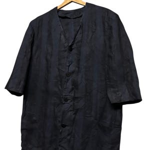 May include: A dark navy blue button-down shirt with a V-neckline and elbow-length sleeves. The garment features a subtle, crinkled texture throughout the fabric and four matching dark buttons down the center front, hanging on a wooden hanger.