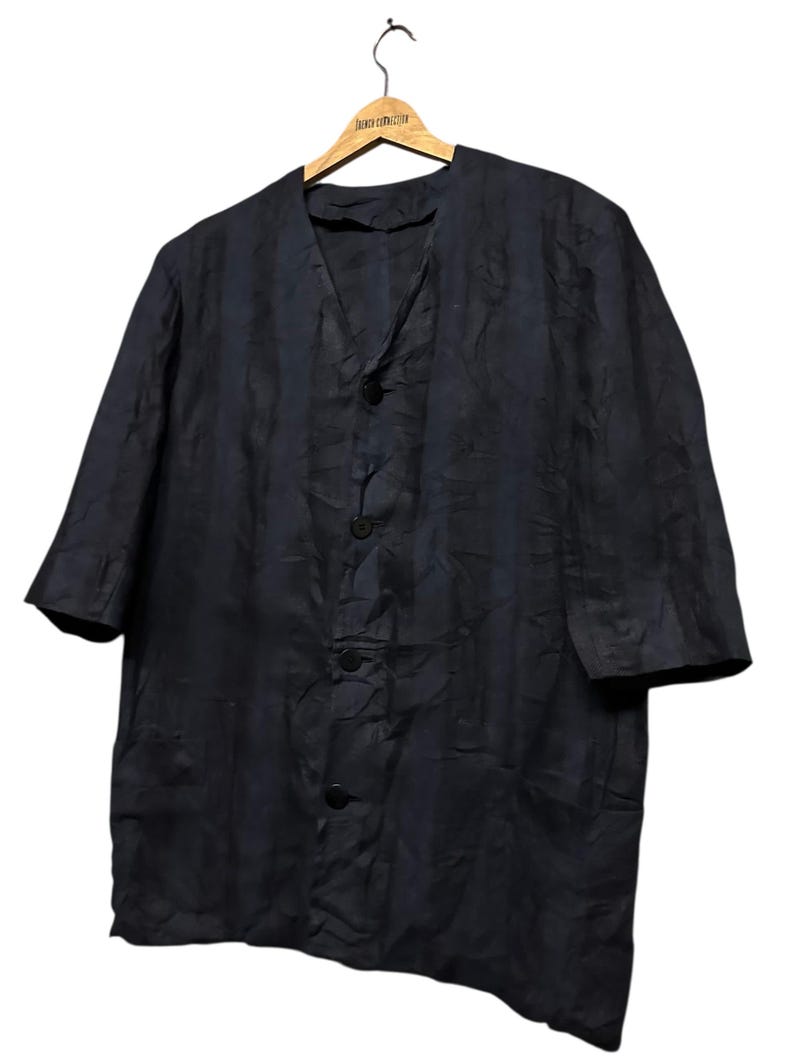 May include: A dark navy blue button-down shirt with a V-neckline and elbow-length sleeves. The garment features a subtle, crinkled texture throughout the fabric and four matching dark buttons down the center front, hanging on a wooden hanger.