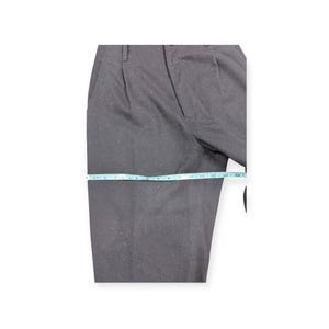 Puede incluir: Pantalones de color gris oscuro con una textura ligeramente moteada y un dise&ntilde;o frontal plisado. Una cinta m&eacute;trica de color azul claro se coloca horizontalmente sobre el muslo, indicando un ancho de unos 35,5 cm en la tela.
