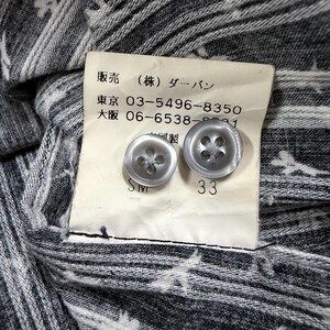 May include: A grey and white patterned fabric garment featuring a cream-colored interior tag with two translucent grey buttons. The tag displays Japanese text, phone numbers 03-5496-8350 and 06-6538-8591, and the number 33 printed below the buttons.