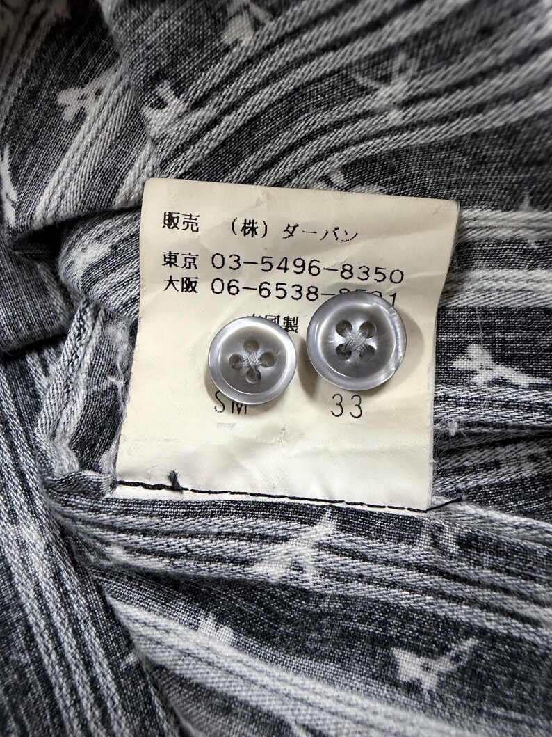 May include: A grey and white patterned fabric garment featuring a cream-colored interior tag with two translucent grey buttons. The tag displays Japanese text, phone numbers 03-5496-8350 and 06-6538-8591, and the number 33 printed below the buttons.