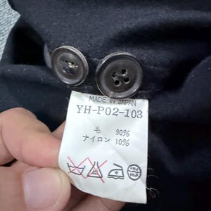 Puede incluir: Una prenda negra con dos botones oscuros y una etiqueta de cuidado blanca. La etiqueta dice MADE IN JAPAN, YH-P02-103, 90% lana y 10% nailon, junto con cuatro s&iacute;mbolos de cuidado est&aacute;ndar, incluyendo no usar lej&iacute;a y limpieza en seco.
