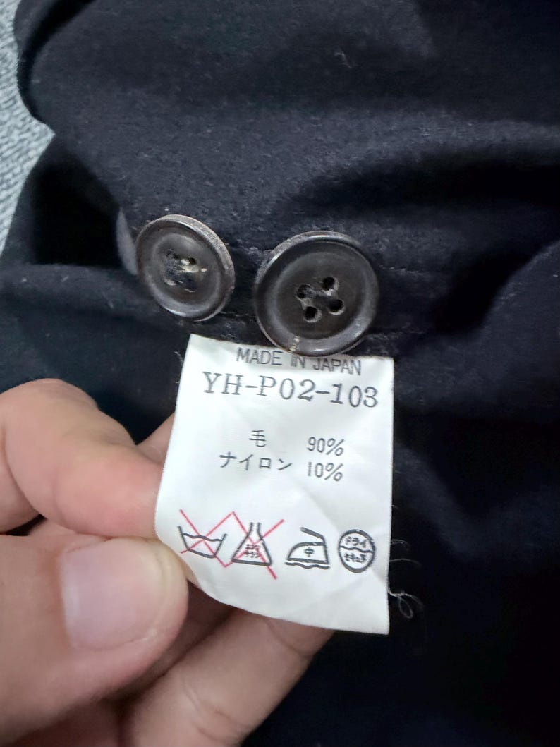 Puede incluir: Una prenda negra con dos botones oscuros y una etiqueta de cuidado blanca. La etiqueta dice MADE IN JAPAN, YH-P02-103, 90% lana y 10% nailon, junto con cuatro s&iacute;mbolos de cuidado est&aacute;ndar, incluyendo no usar lej&iacute;a y limpieza en seco.