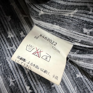 May include: A white fabric care label attached to a dark gray and white striped garment. The label features the text X462022, 100% cotton in Japanese, and laundry symbols including a hand wash icon, a no-bleach symbol, and a high-heat iron icon.