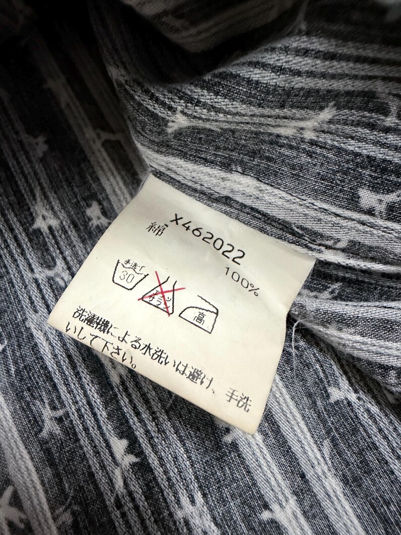 May include: A white fabric care label attached to a dark gray and white striped garment. The label features the text X462022, 100% cotton in Japanese, and laundry symbols including a hand wash icon, a no-bleach symbol, and a high-heat iron icon.