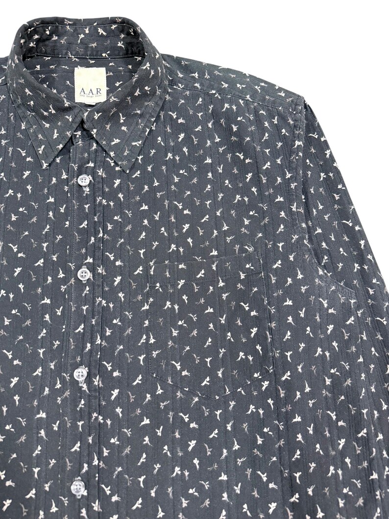 May include: A dark charcoal gray long-sleeve button-down shirt featuring a repeating white floral pattern and vertical pinstripes. The shirt includes a standard collar, a front chest pocket, and a label at the neck with the text A.A.R.
