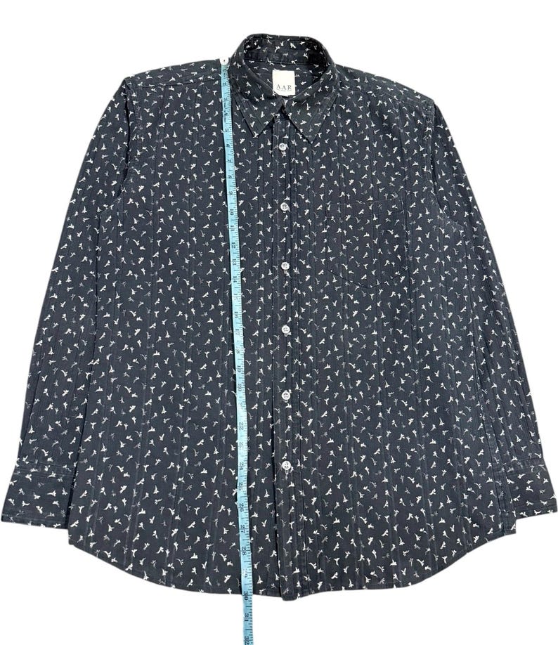 May include: A dark gray long-sleeve button-down shirt with a white repeating pattern of small figures. A blue measuring tape is placed vertically along the center, showing a length of approximately 30 inches from the collar to the bottom hem.