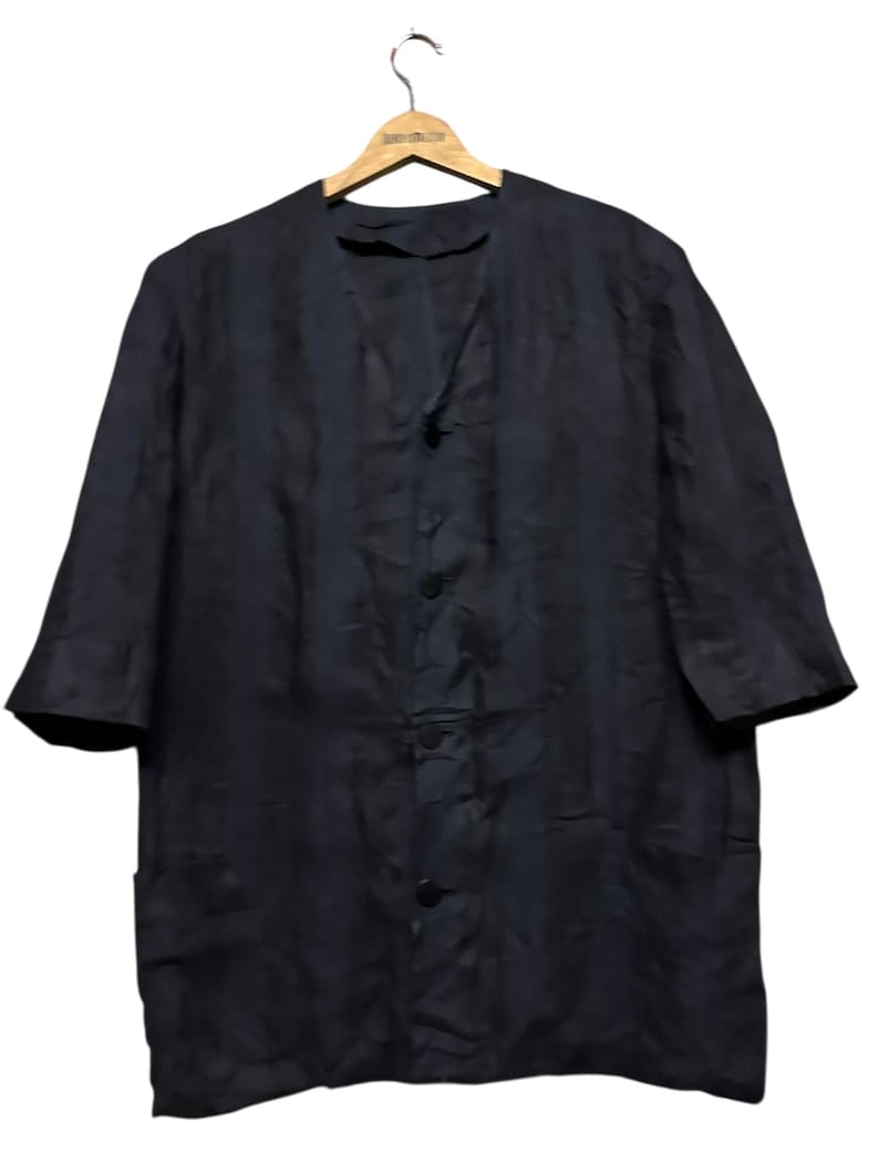 May include: A dark navy blue button-down shirt with a subtle vertical striped pattern, featuring a collarless V-neckline, three-quarter length sleeves, and two front pockets, displayed on a wooden hanger against a plain background.