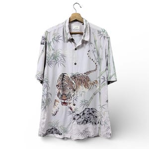 Può includere: Una camicia a maniche corte color grigio chiaro con abbottonatura frontale, caratterizzata da una stampa con una tigre che salta, steli di bambù verdi e formazioni rocciose grigie. La camicia ha un colletto classico ed è su una gruccia.
