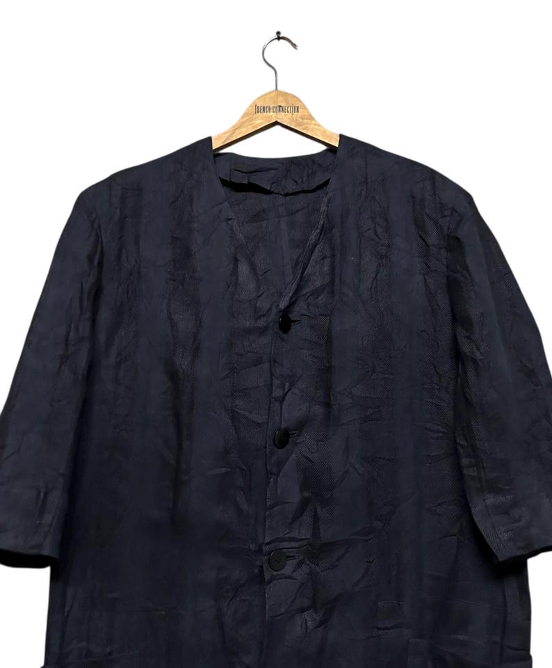 May include: A dark navy blue jacket with a collarless V-neckline and three black buttons down the front. The garment features a textured, slightly wrinkled fabric finish and three-quarter length sleeves, hanging on a wooden clothes hanger.