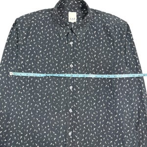 May include: A dark gray long-sleeve button-down shirt with a white floral pattern and vertical textured stripes. A light blue measuring tape is placed across the chest, showing a width of approximately 22 inches. The shirt features a collar and a pocket.