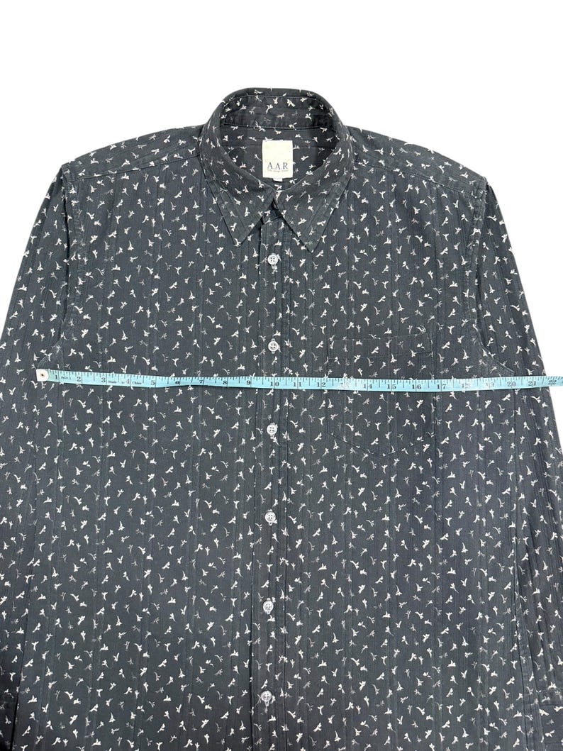 May include: A dark gray long-sleeve button-down shirt with a white floral pattern and vertical textured stripes. A light blue measuring tape is placed across the chest, showing a width of approximately 22 inches. The shirt features a collar and a pocket.