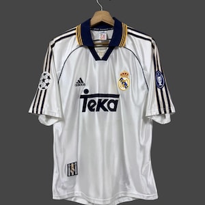 May include: A white short-sleeved soccer jersey with a navy blue collar featuring yellow trim. The shirt displays the Adidas logo, the Teka sponsor text, and the Real Madrid crest. It includes Champions League sleeve patches and a small emblem.