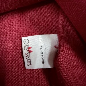 Puede incluir: Primer plano de una tela roja con una etiqueta blanca adjunta. La etiqueta tiene instrucciones de lavado y texto japon&eacute;s. La tela parece ser un material tejido, posiblemente lino o algod&oacute;n.