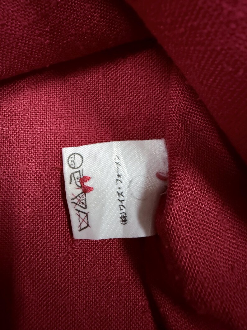 Puede incluir: Primer plano de una tela roja con una etiqueta blanca adjunta. La etiqueta tiene instrucciones de lavado y texto japon&eacute;s. La tela parece ser un material tejido, posiblemente lino o algod&oacute;n.