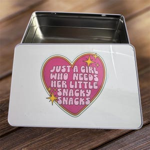 May include: A rectangular metal tin with an open hinged lid. The lid displays a pink heart design with the text "JUST A GIRL WHO NEEDS HER LITTLE SNACKY SNACKS" in a retro font, with yellow star accents.