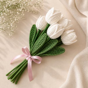 Puede incluir: Un ramo de tulipanes blancos de ganchillo con tallos y hojas verdes, atado con una cinta de sat&eacute;n rosa claro. Las flores presentan una construcci&oacute;n de hilo texturizado y los tallos son largos y rectos, creando un arreglo floral estructurado.