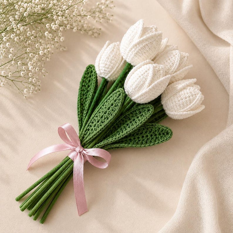 Puede incluir: Un ramo de tulipanes blancos de ganchillo con tallos y hojas verdes, atado con una cinta de sat&eacute;n rosa claro. Las flores presentan una construcci&oacute;n de hilo texturizado y los tallos son largos y rectos, creando un arreglo floral estructurado.