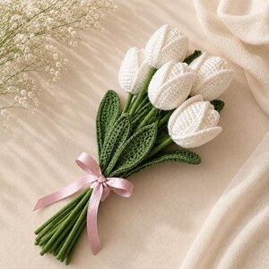Puede incluir: Un ramo de cinco tulipanes de ganchillo blancos con tallos y hojas de ganchillo verdes. Los tallos est&aacute;n agrupados y atados con una cinta de sat&eacute;n rosa claro. La textura del hilo es visible en los p&eacute;talos y en el follaje.