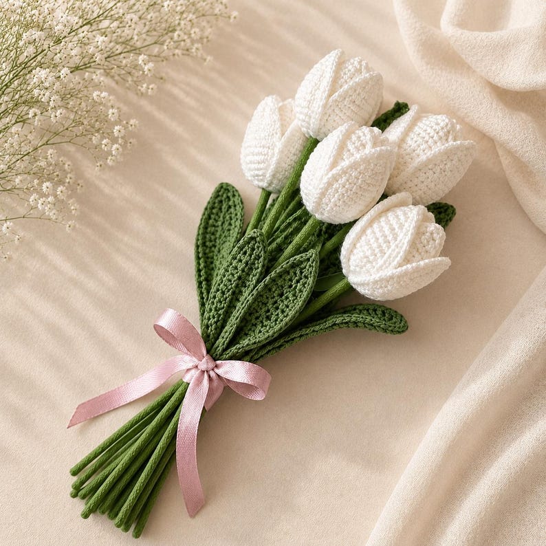 Puede incluir: Un ramo de cinco tulipanes de ganchillo blancos con tallos y hojas de ganchillo verdes. Los tallos est&aacute;n agrupados y atados con una cinta de sat&eacute;n rosa claro. La textura del hilo es visible en los p&eacute;talos y en el follaje.
