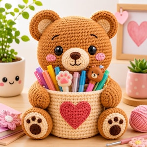 Peut inclure: Un organisateur de bureau en forme d'ours en crochet marron, doté d'un panier cylindrique couleur crème orné d'un grand cœur rouge. L'ours a des yeux noirs, des joues roses et des coussinets sur les pattes. Il contient divers stylos colorés.