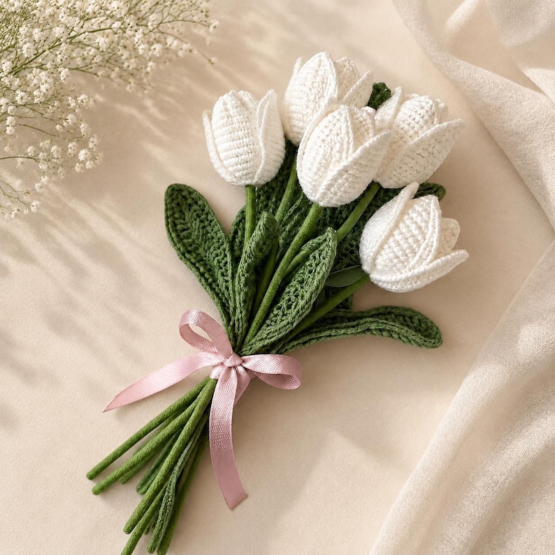 Puede incluir: Un ramo de cinco tulipanes blancos tejidos a ganchillo con tallos y hojas verdes tambi&eacute;n tejidos, atados con una cinta de sat&eacute;n rosa claro. Los tulipanes presentan una textura de hilo y est&aacute;n dispuestos en un grupo con tallos largos.