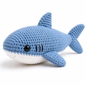 May include: A blue and white crocheted shark plush toy featuring a textured yarn surface, a single dorsal fin, two side fins, a forked tail, and a black plastic eye. The shark has three small black embroidered gill slits on its side.