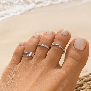Può includere: Un set di tre anelli da dito del piede color argento indossati sulle dita. Un anello presenta un design a bande multiple, un altro è una fascia sottile incastonata con piccole pietre chiare e il terzo è una fascia in metallo intrecciato.