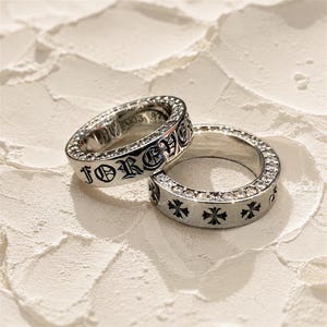 May include: Two silver rings with detailed designs. One ring has the word "FOREVER" in gothic script and is set with clear gemstones. The other ring features a pattern of cross symbols and is also embellished with gemstones.