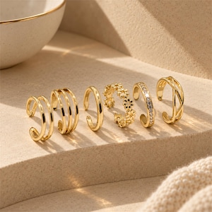 May include: A set of six gold-toned ear cuffs featuring various designs, including a double band, a triple band, a plain band, a floral-shaped band, a band encrusted with clear stones, and an openwork crisscross band, all with an open design.