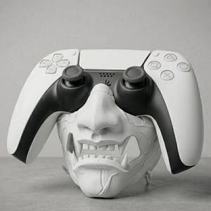 Peut inclure: Une manette de jeu vidéo blanche et noire posée sur un masque sculpté blanc représentant un visage stylisé avec un nez et des dents saillantes. La manette comprend des boutons directionnels, deux joysticks et un pavé tactile central.